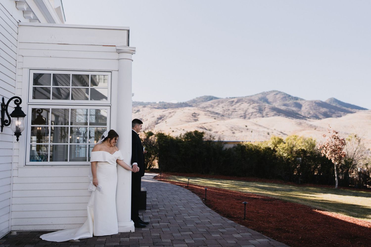 Bride and groom share a first look moment beside a white building with scenic mountain views in the background.