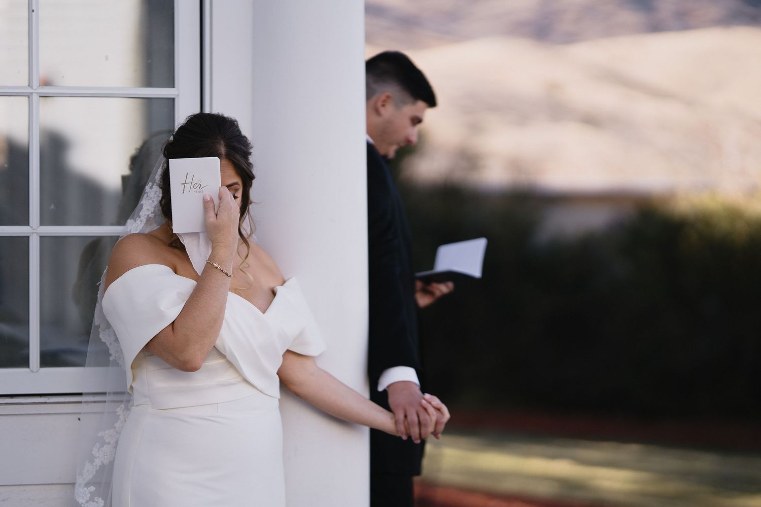 Bride and groom share private vows around a column, bride holding letter to face in white gown and veil.
