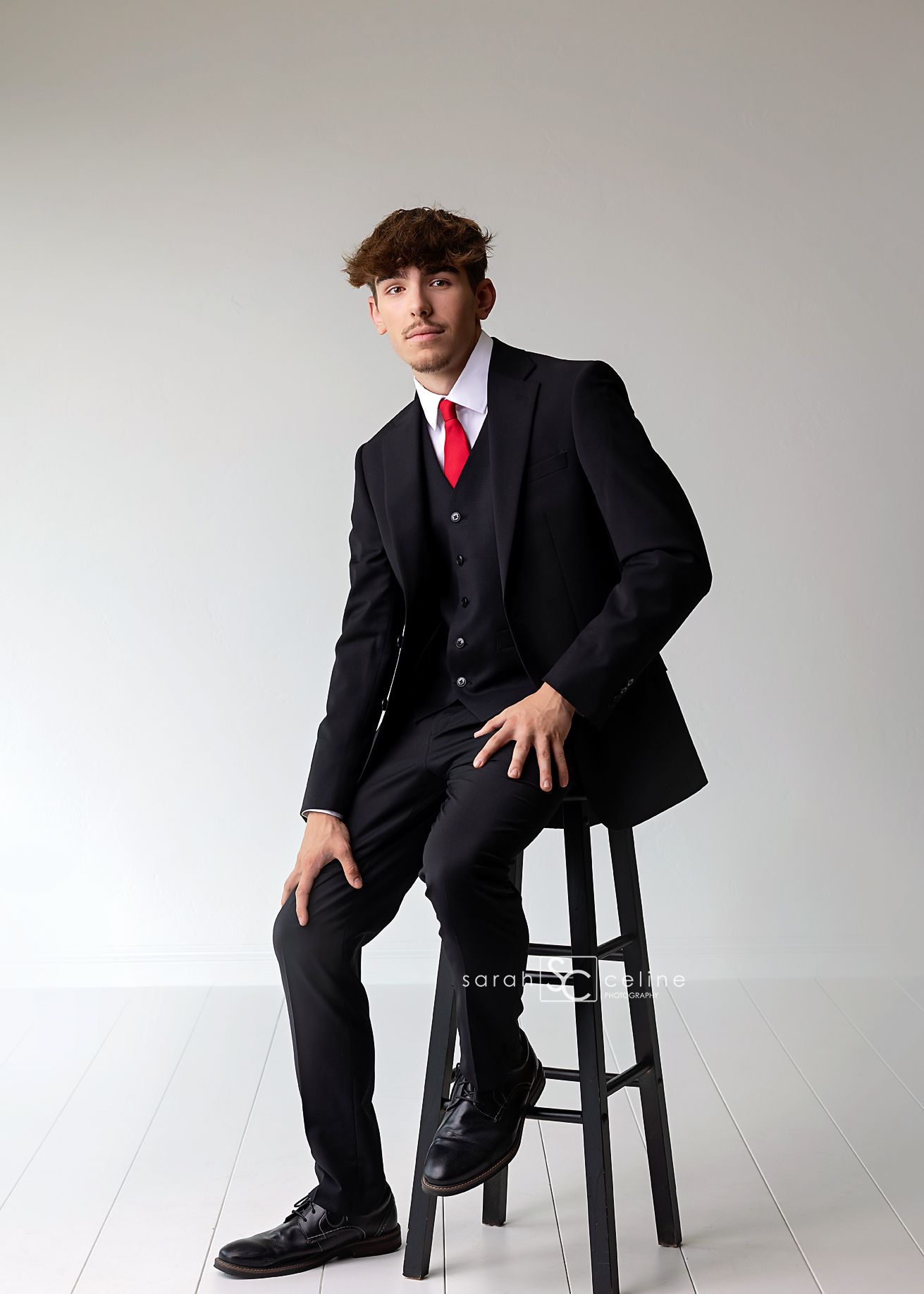 Young man in black suit with red tie seated on a stool, posing for a formal portrait photo.