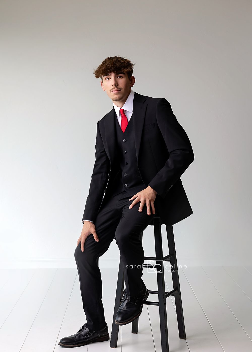 Young man in black suit with red tie seated on a stool, posing for a formal portrait photo.