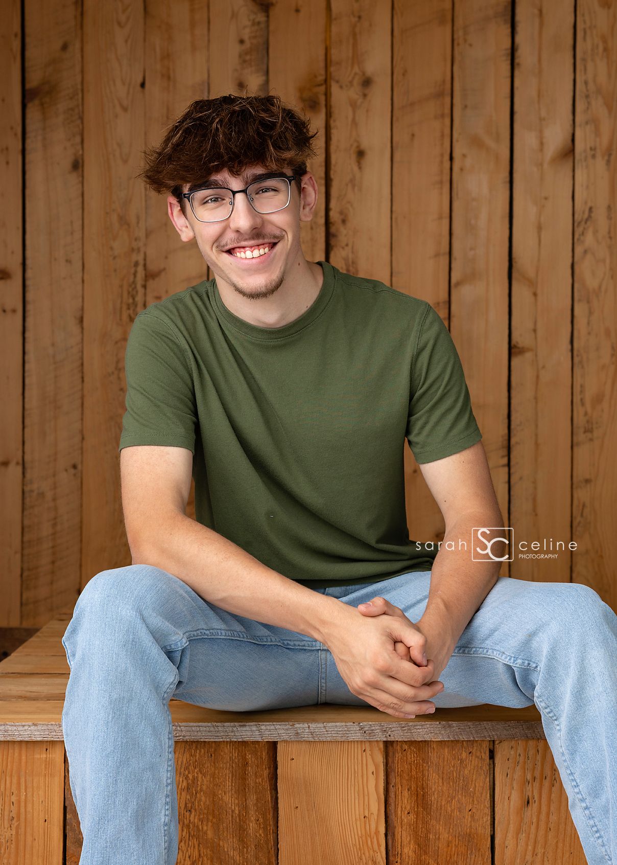 Smiling young man with glasses wearing a green t-shirt and jeans, sitting on a wooden bench against a wood panel wall.