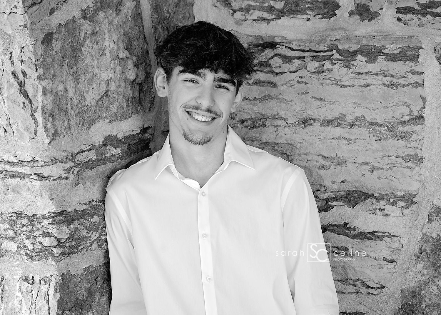 Black and white portrait of a smiling young man in a white shirt leaning against a rustic stone wall.