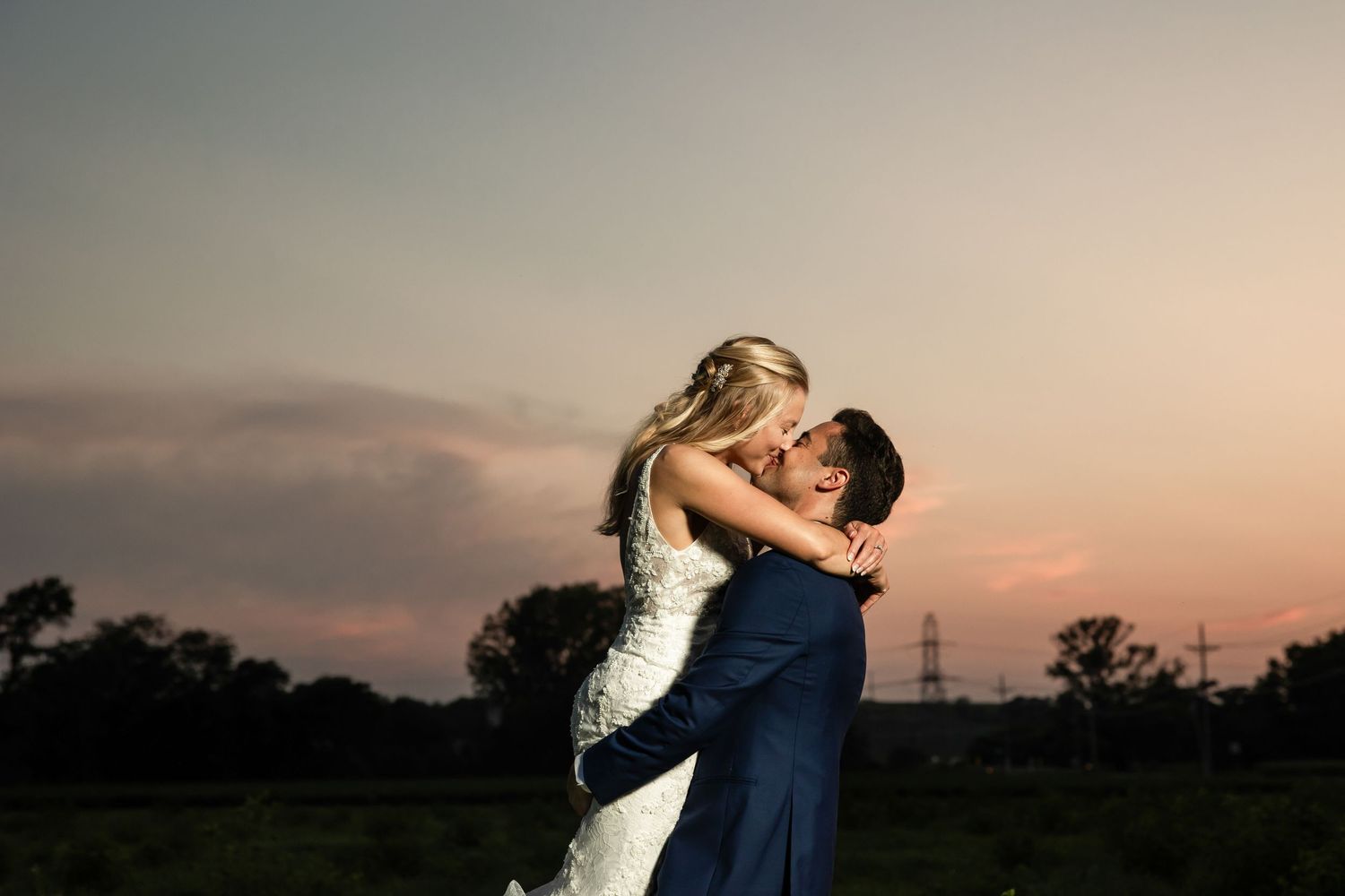 Couple sharing a romantic kiss at sunset, groom lifting bride in wedding attire against a colorful dusk sky.