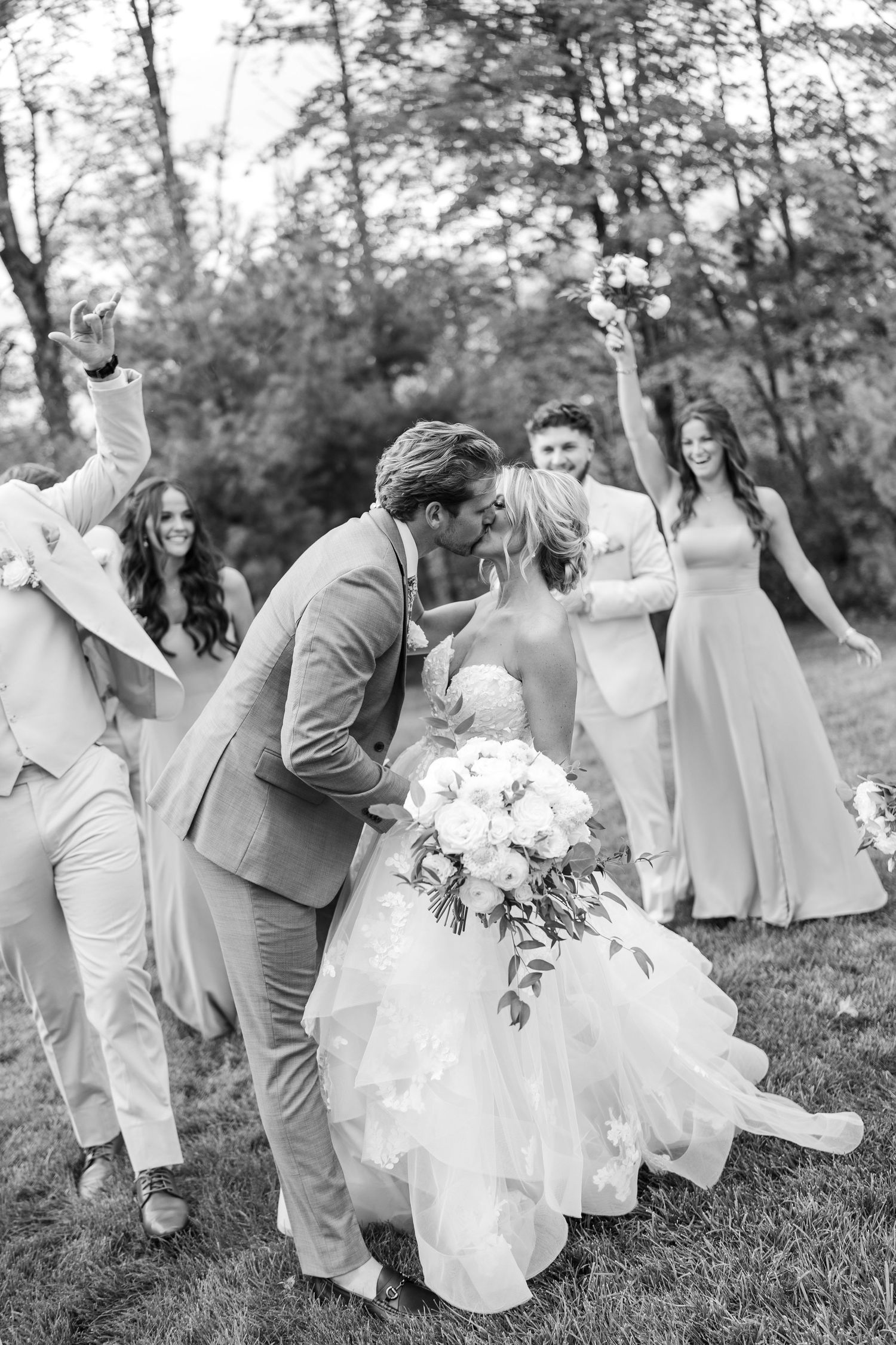 Black and white photo of a bride and groom kissing surrounded by cheering wedding party outdoors.