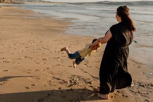 Mother in black dress swinging her child on a sandy beach at sunset.