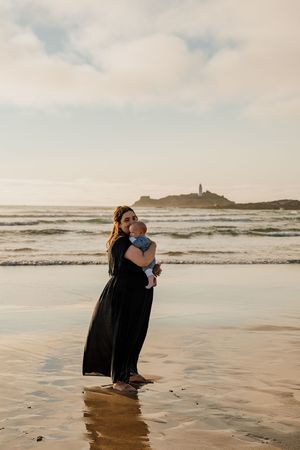 Woman in black dress holds baby on sandy beach at sunset with ocean waves and lighthouse in background.