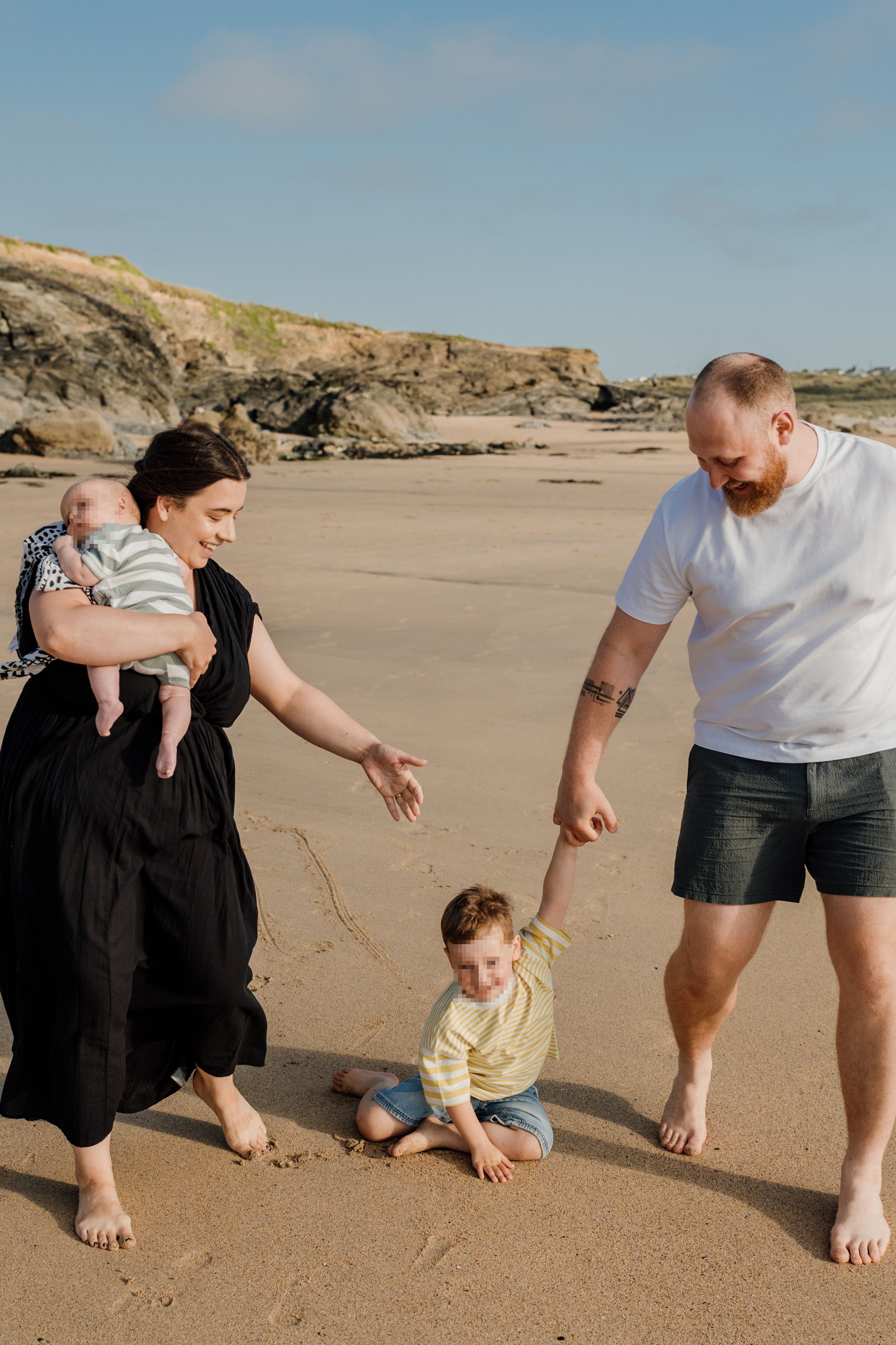 Family of four enjoying a sunny beach day, with mother holding baby and father helping toddler on sand.