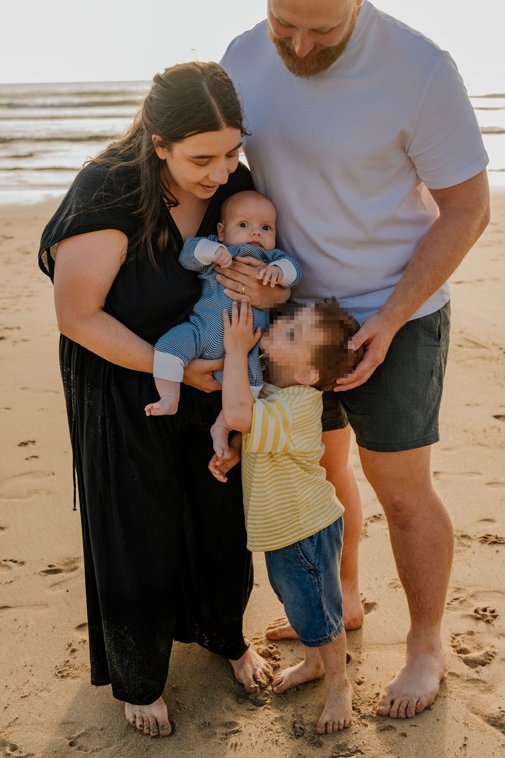 Family of four with a baby and toddler sharing a warm moment together on a sandy beach at sunset.