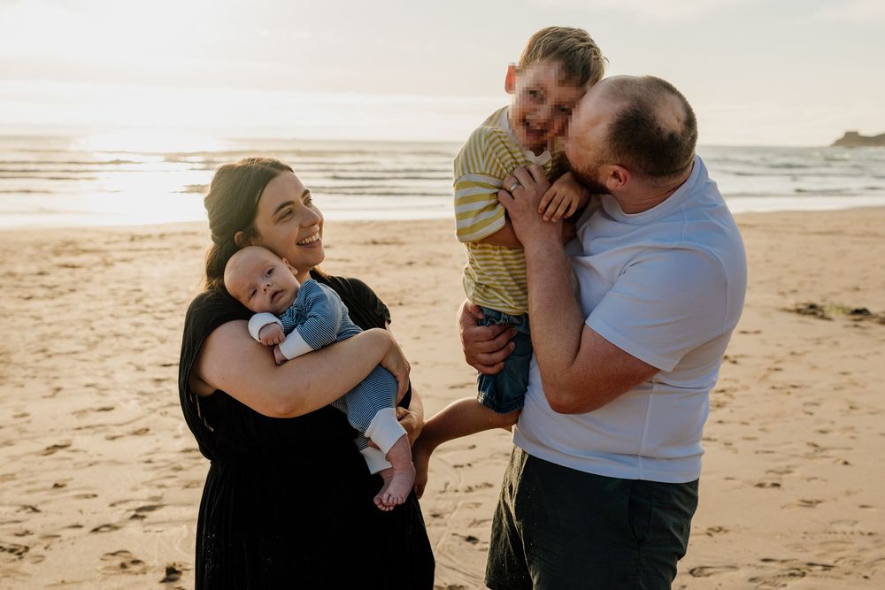 Family of four enjoying a sunset beach day, parents holding baby and young boy, smiling and laughing together.
