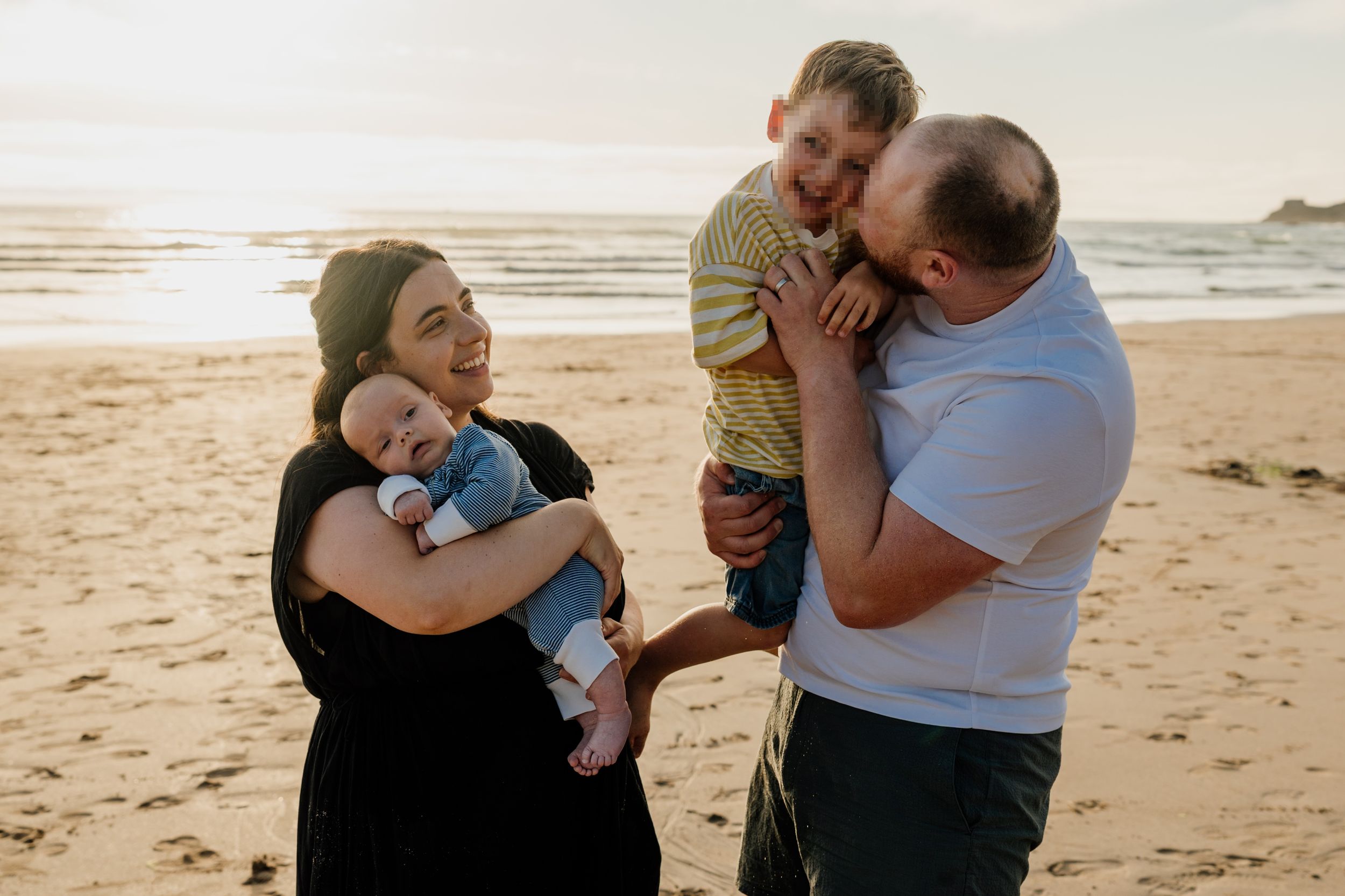 Family of four enjoying a sunset beach day, parents holding baby and young boy, smiling and laughing together.