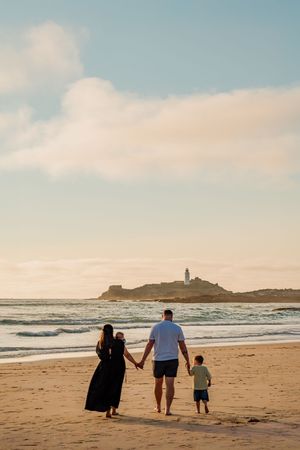 Family of four walking hand-in-hand on a sandy beach at sunset with a lighthouse on rocky headland in background.