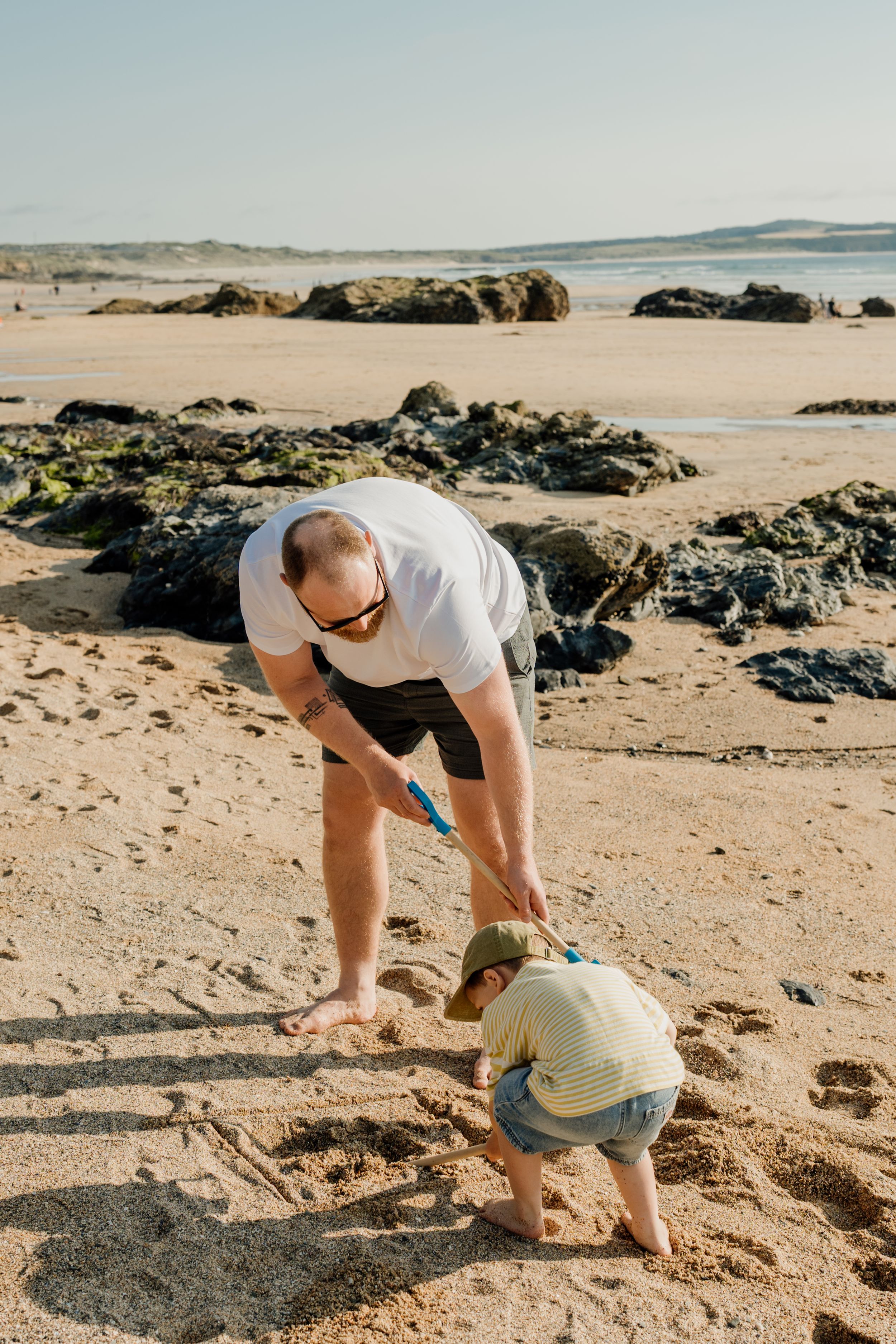 Father and toddler playing in the sand on a rocky beach, digging together with blue shovels on a sunny day.