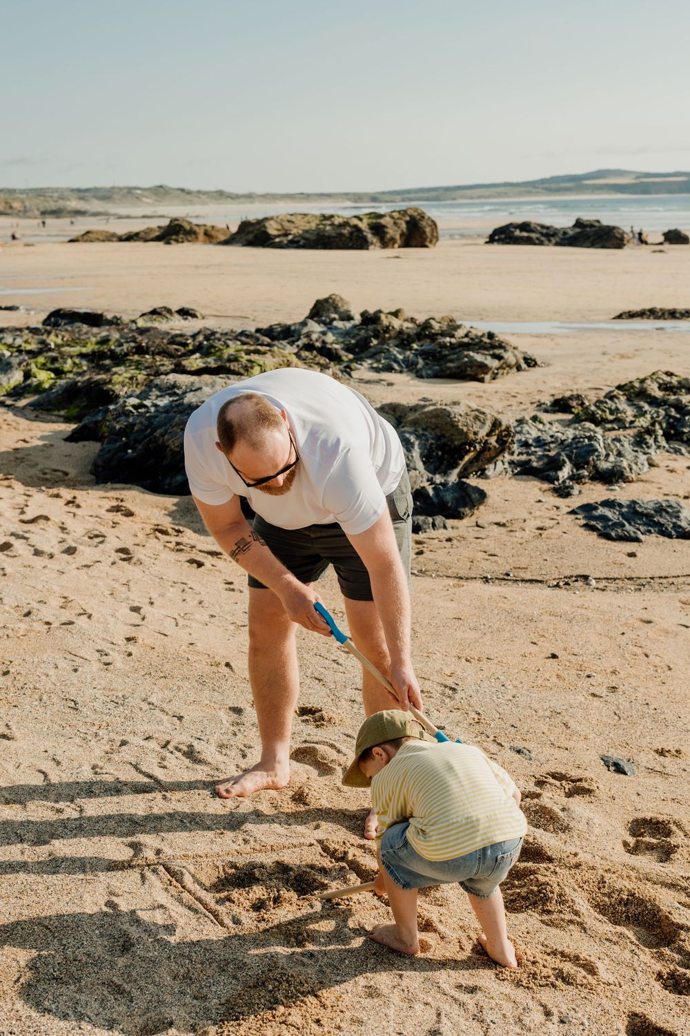 Father and toddler playing in the sand on a rocky beach, digging together with blue shovels on a sunny day.