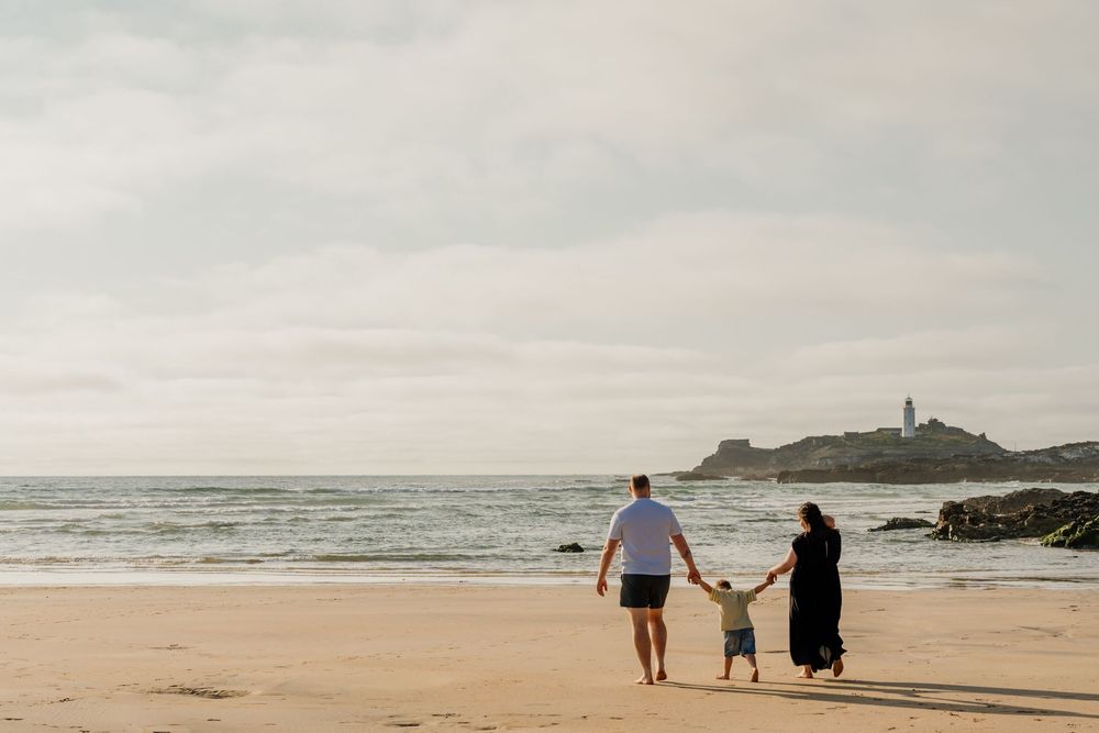 Family of three walking hand-in-hand on a sandy beach with a lighthouse in the background on an overcast day.