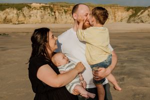 Family of four enjoying a sunny beach day, parents holding two young children with sandy cliffs in the background.