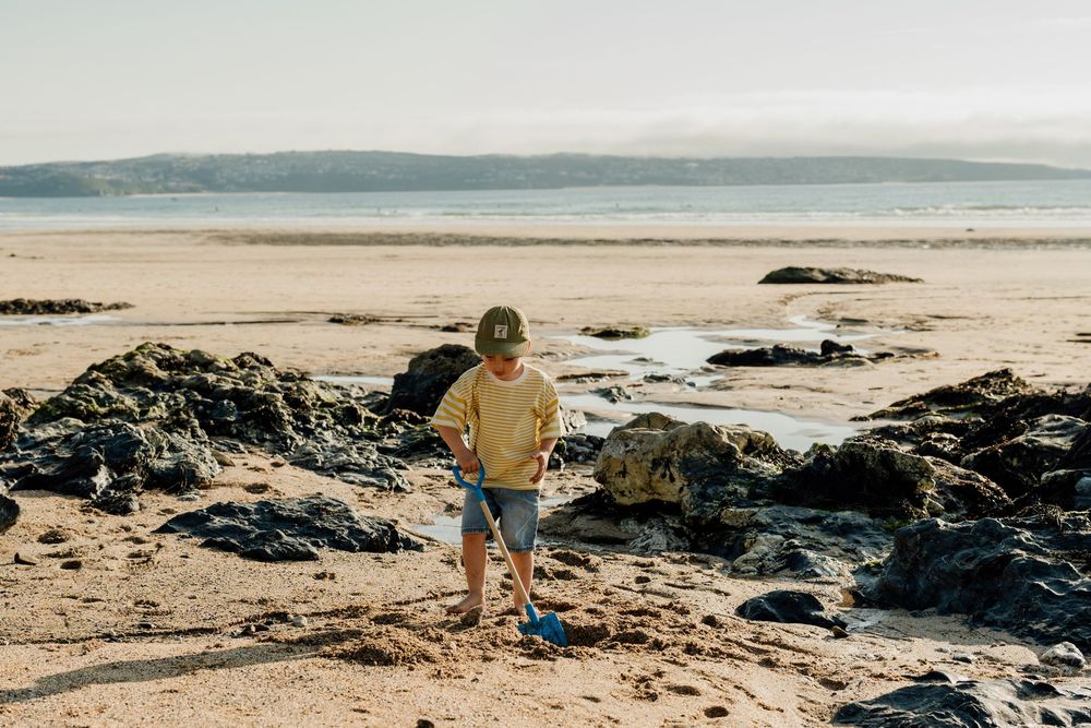A young boy playing on a sandy beach with a blue shovel, surrounded by rocks with mountains in the background.