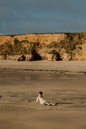 Child sitting alone on sandy beach with dramatic golden cliffs and dark sky in background.