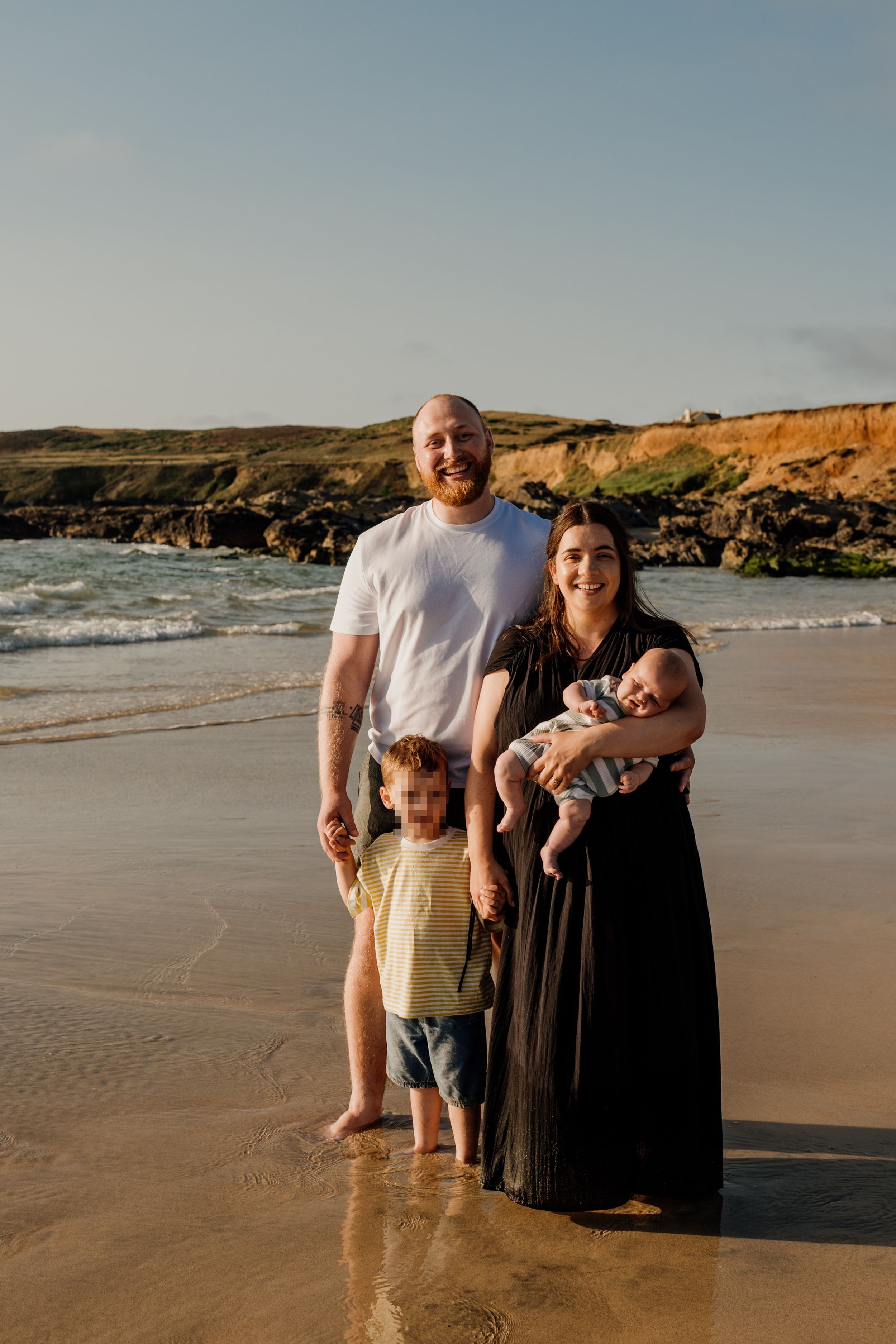 Family of four posing on a sandy beach at golden hour, with ocean waves and coastal cliffs in the background.