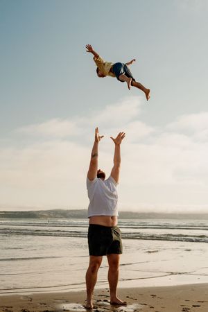 A man tosses a child high into the air on a sunny beach, with ocean waves and blue sky in the background.