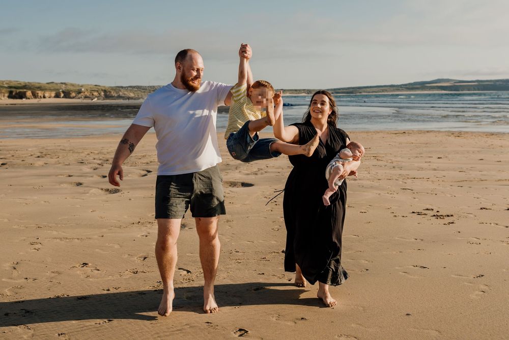 Family of four enjoying a sunny beach walk, dad lifting toddler overhead, mom cradling newborn, sandy shore backdrop.