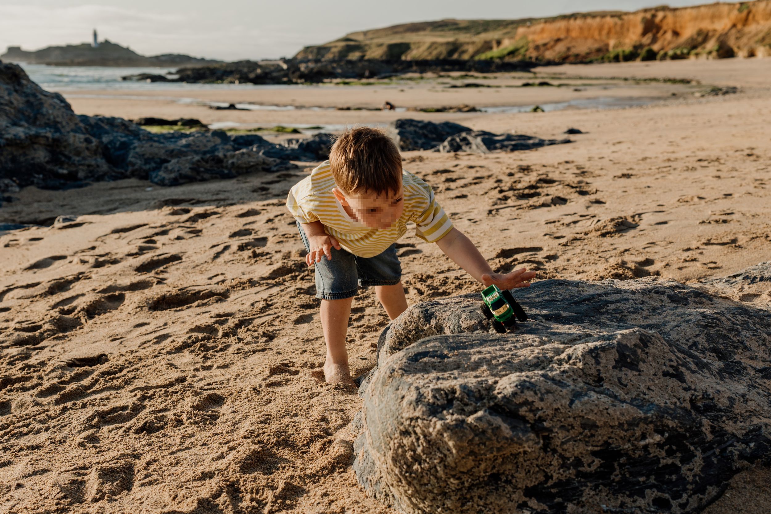 Young boy climbing a large rock on a sandy beach with rocky cliffs and a lighthouse in the background.