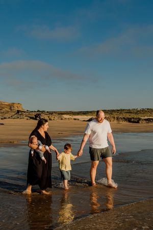 Family of four walking through shallow beach water at golden hour, parents holding hands with toddler.