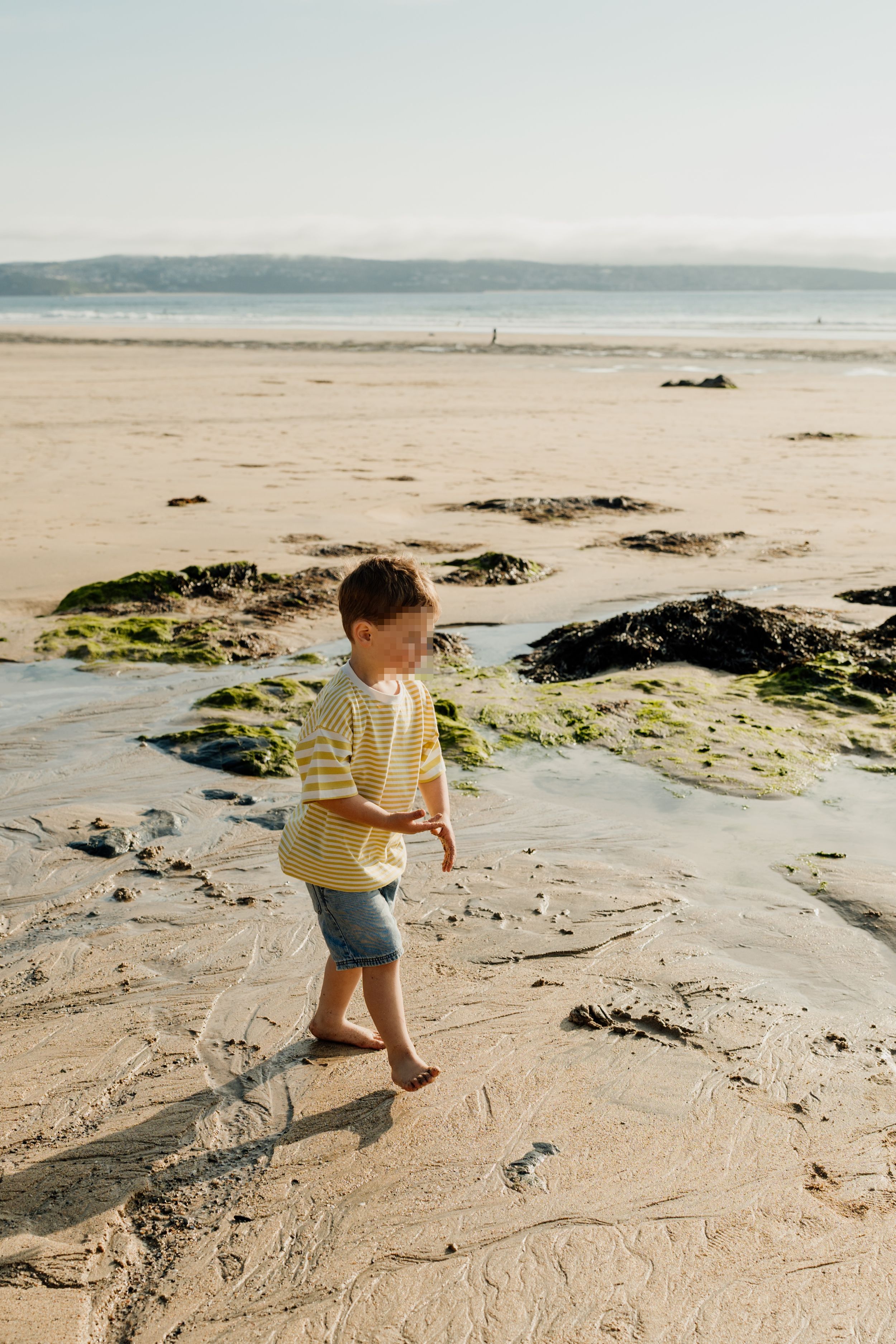 A young boy in a striped shirt and denim shorts walks barefoot on a sandy beach with rocks covered in green algae.