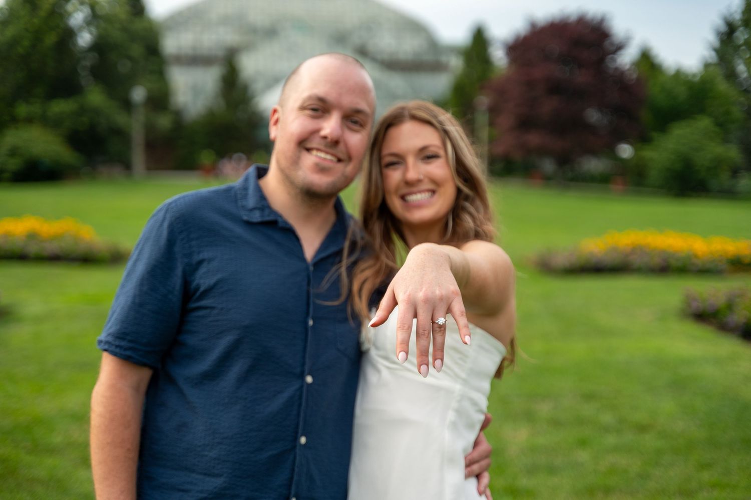 Happy couple smiling outdoors in a lush green park, woman showing off engagement ring on her hand.