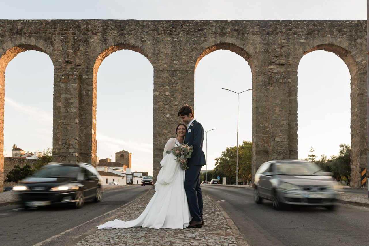 A romantic wedding photo taken between stone arches of an aqueduct with blurred cars passing on both sides at dusk.