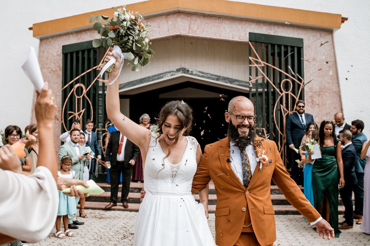Joyous newlyweds celebrate outside church with guests, bride in white dress and groom wearing distinctive rust-colored suit.