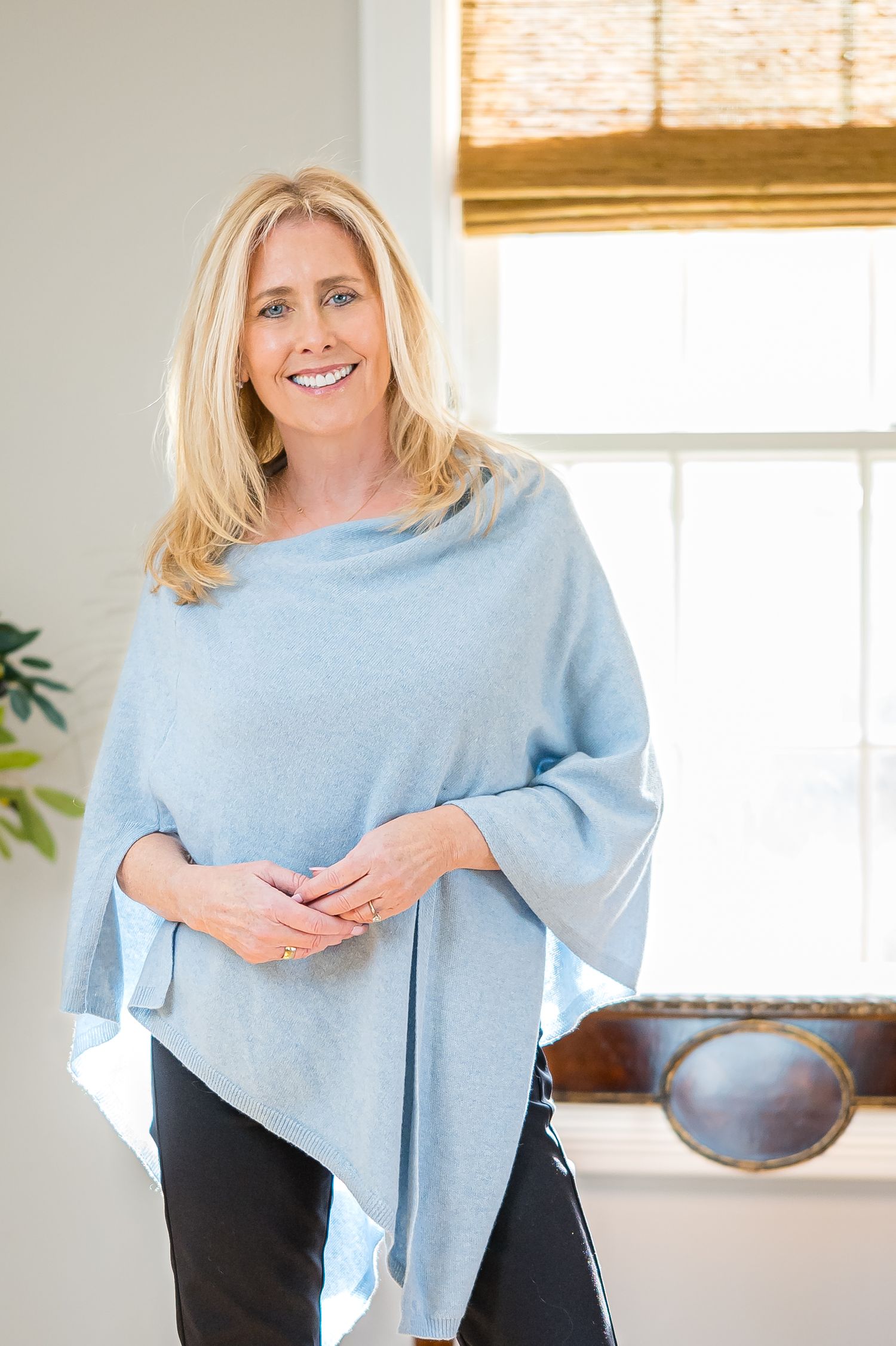 Smiling blonde woman wearing a light blue asymmetric knit poncho top with black pants, standing indoors near a bright window.