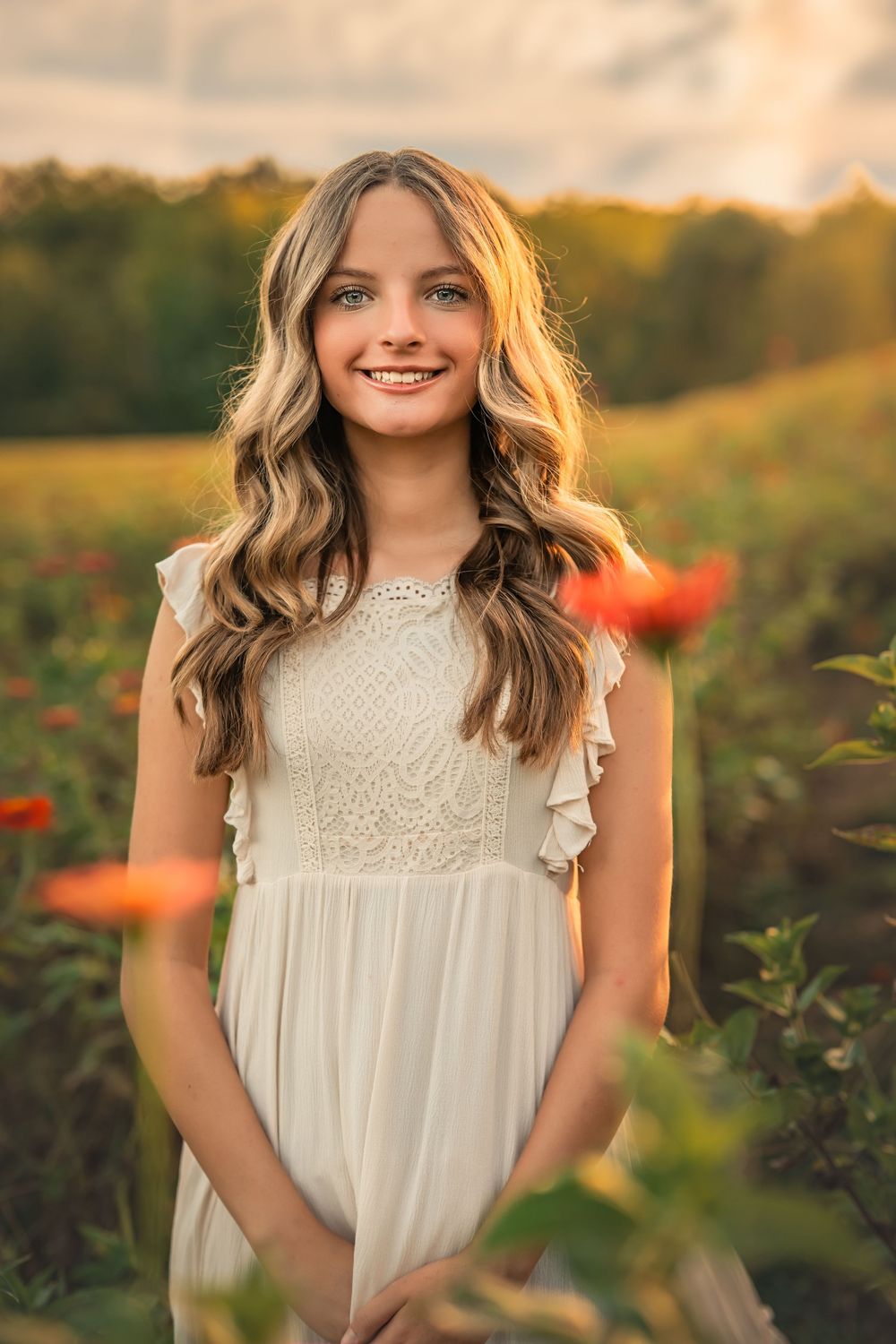 Smiling teenage girl with long wavy hair in a white dress standing in a colorful flower field at sunset.
