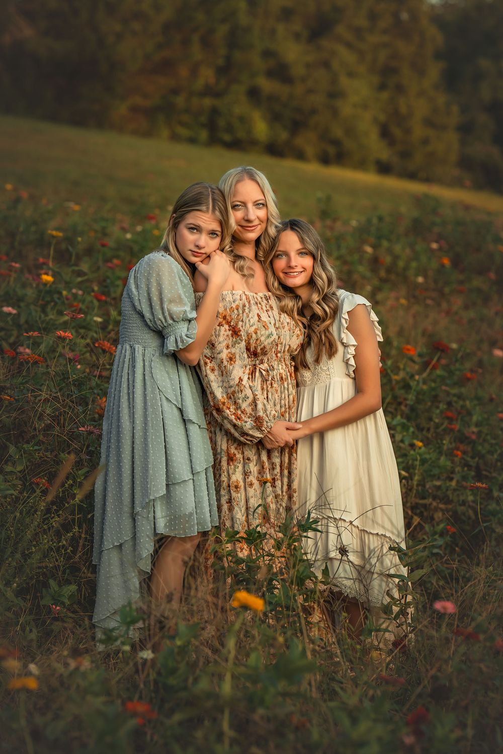 Pregnant woman standing with two daughters in a sunlit flower field, all wearing elegant dresses.