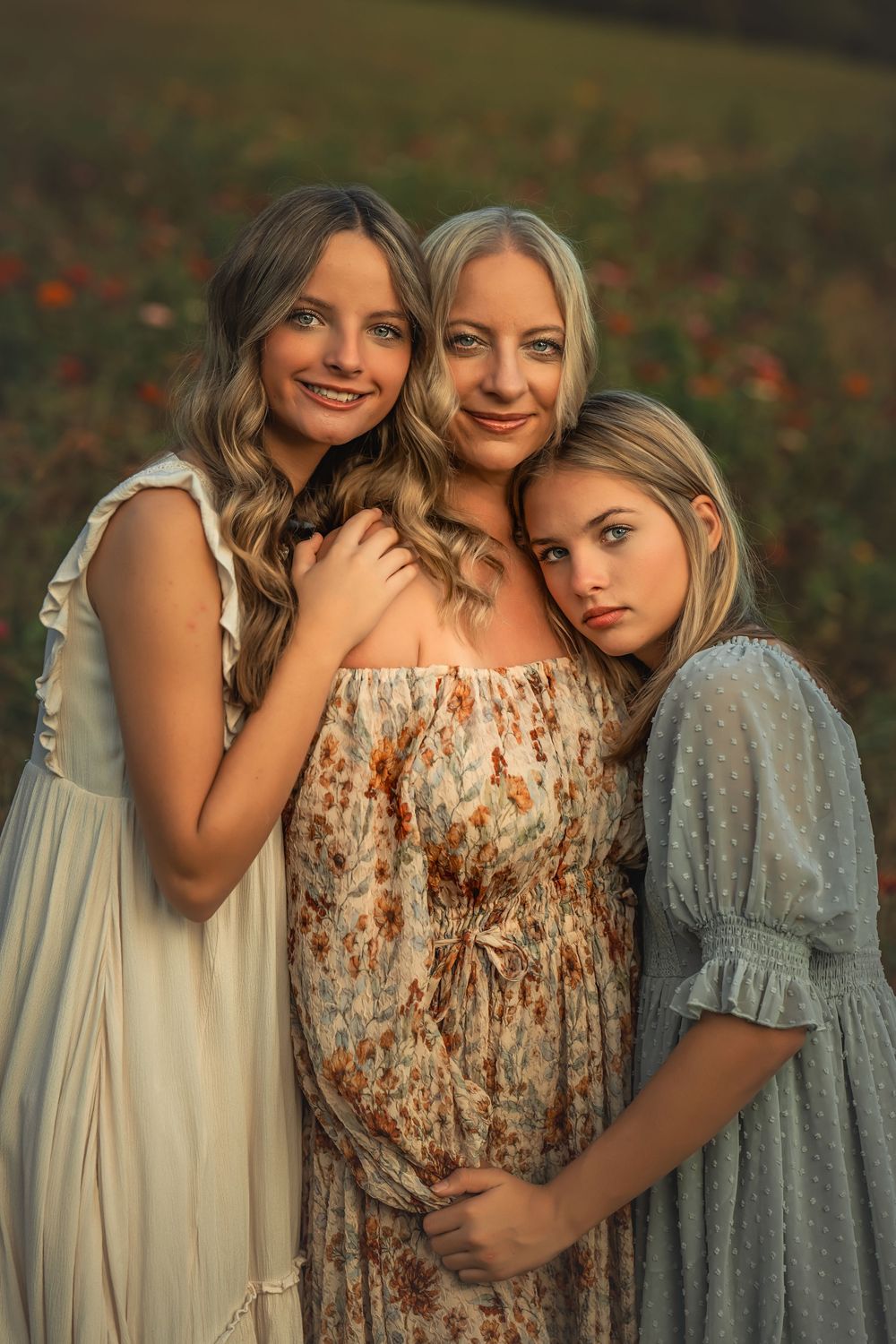 Pregnant blonde mother posing with two daughters in flowing dresses in a vibrant flower field.
