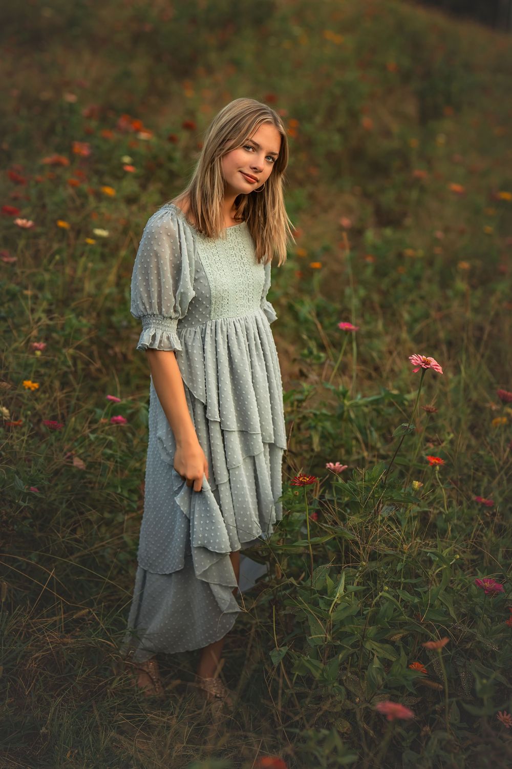 A young blonde girl in a light blue tiered dress stands in a colorful wildflower field at golden hour.