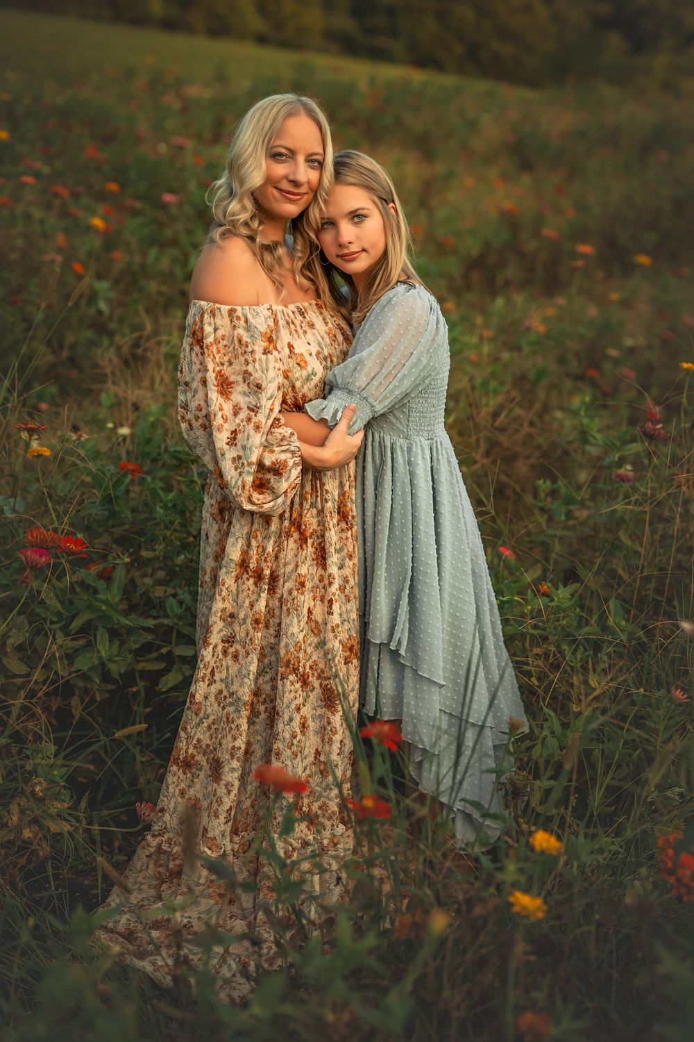 Mother and daughter in floral and blue dresses embrace in a sunlit wildflower field during golden hour.