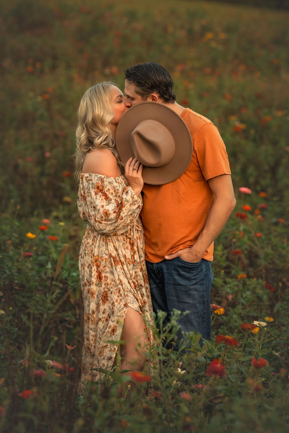Couple sharing a kiss hidden behind a brown hat in a blooming flower field at dusk.