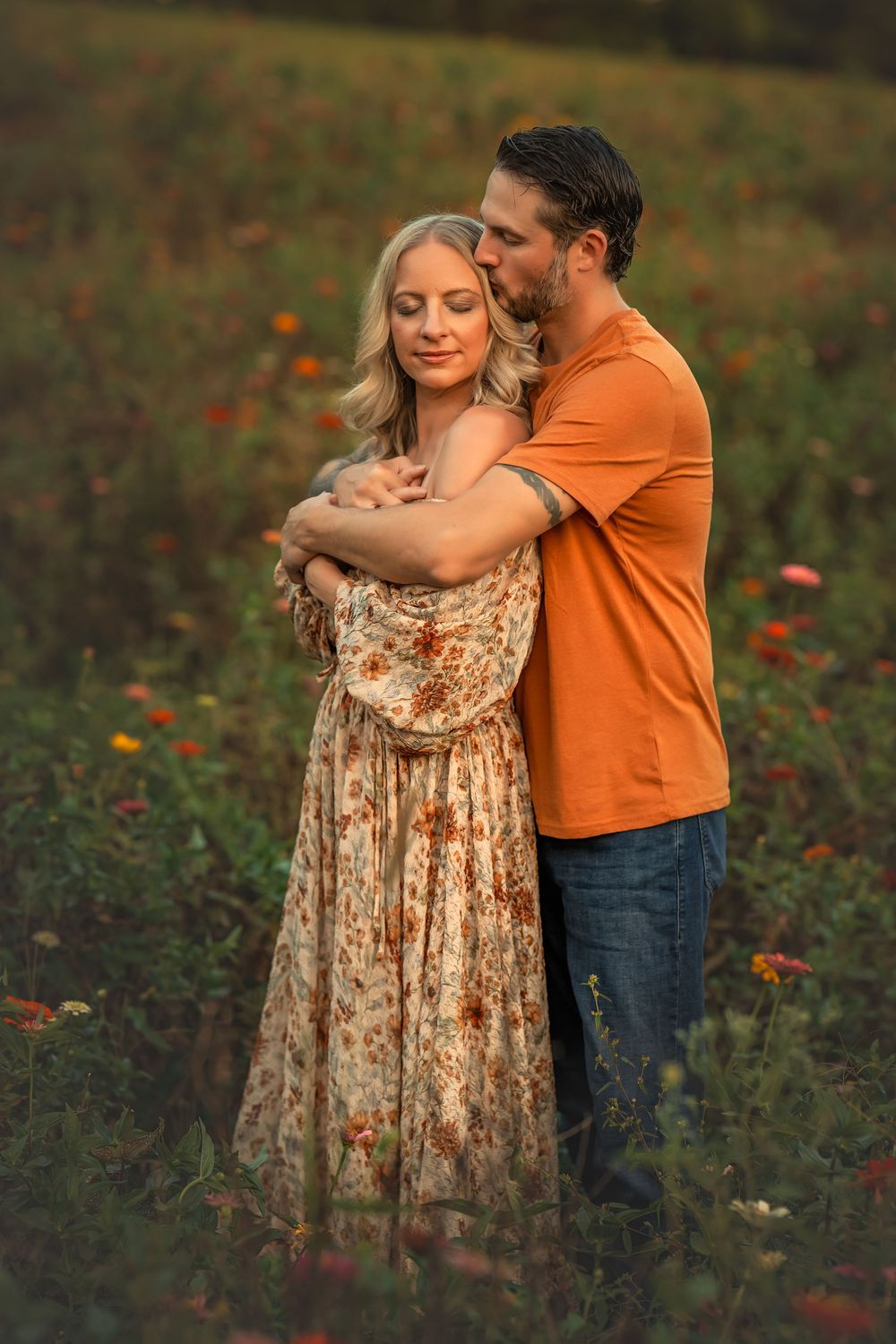 Couple embracing in a flower field, woman in floral dress, man in orange shirt, golden hour lighting.