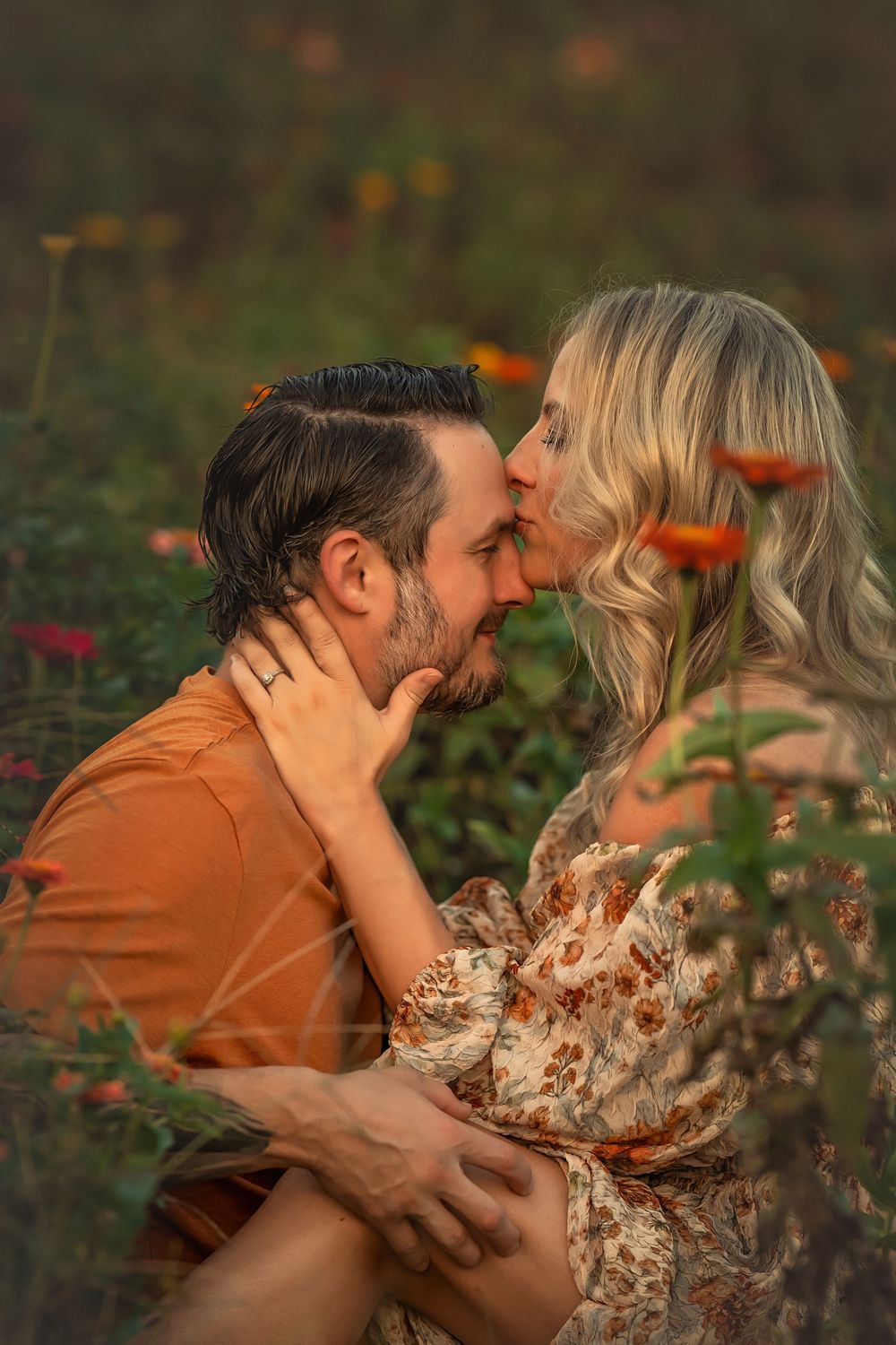 A couple shares a romantic forehead-kiss moment sitting in a colorful flower field at golden hour.