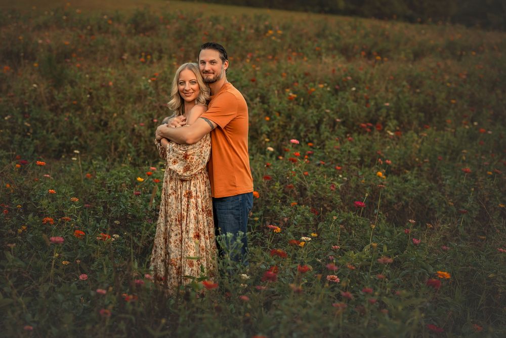 Couple embracing in a colorful wildflower field at golden hour, man in orange shirt hugging woman in floral dress.