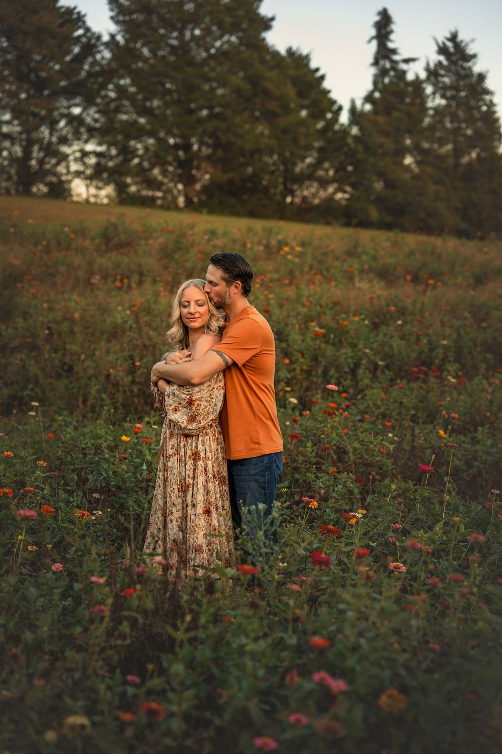 Couple embracing in a colorful flower field at golden hour, woman in floral dress, man in orange shirt.
