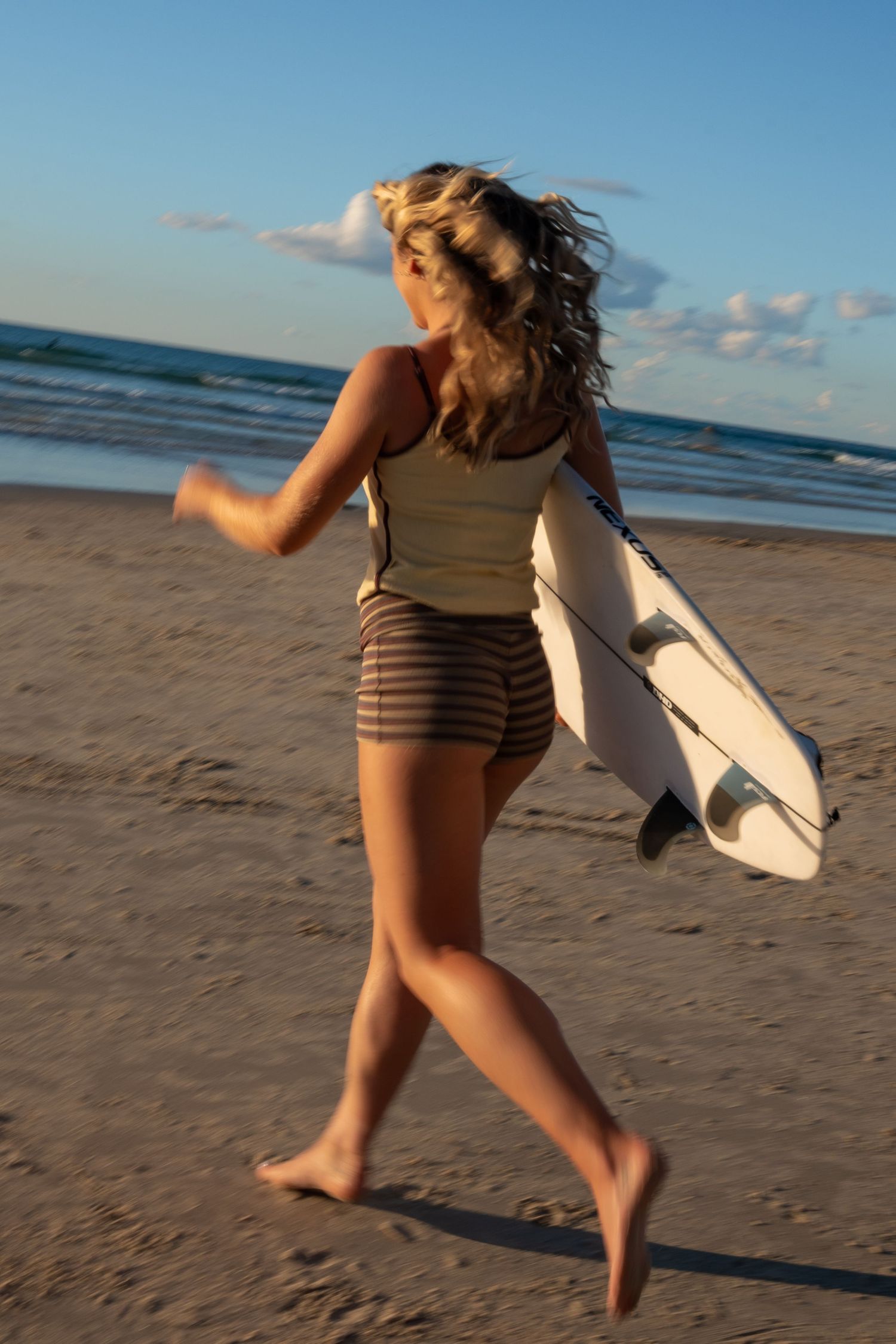 A blonde woman in striped shorts runs barefoot on a sandy beach carrying a white surfboard at sunset.