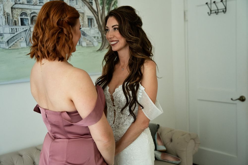 Bride in lace wedding gown smiling with bridesmaid in mauve dress during wedding preparations indoors.