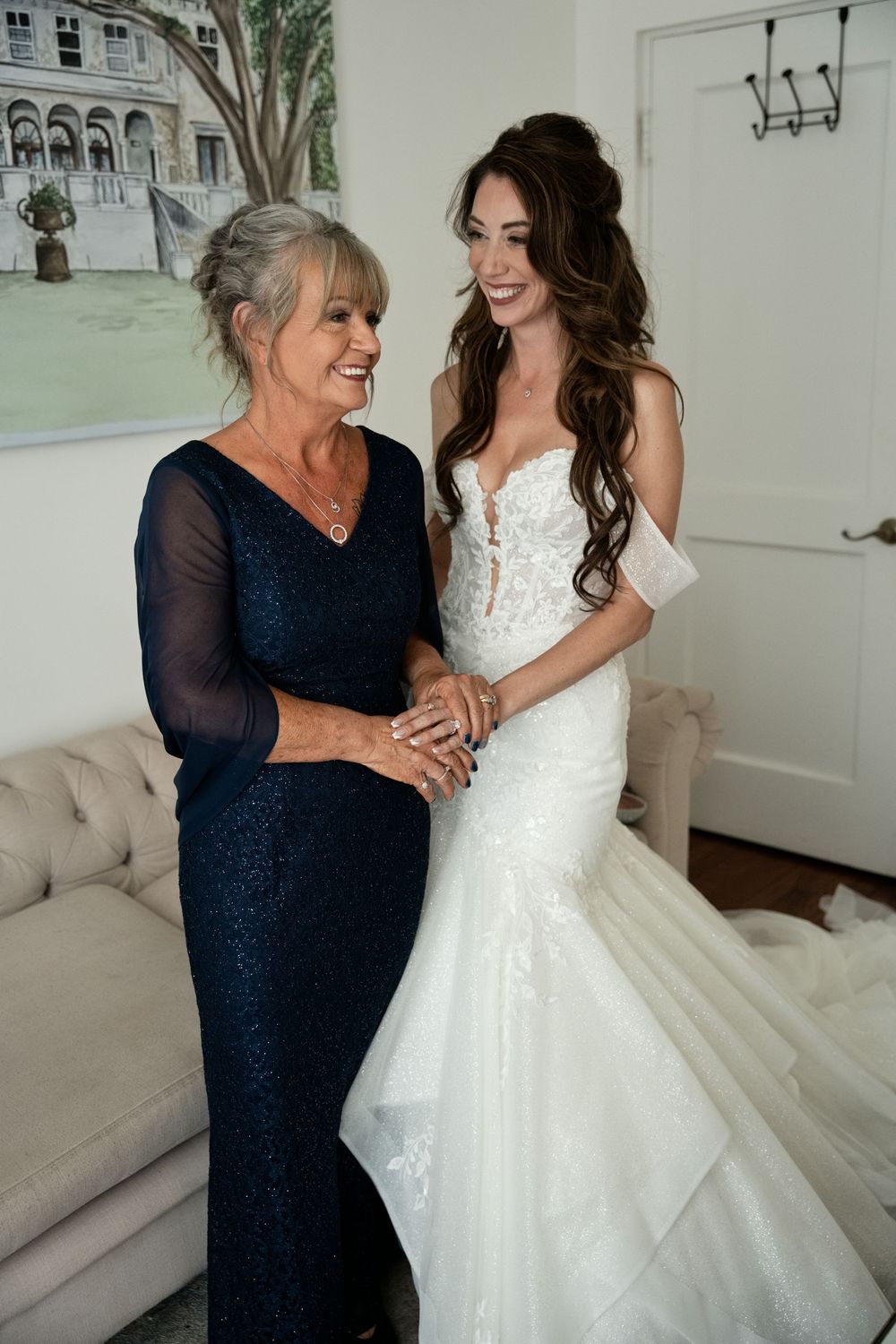 Bride in white mermaid gown smiles with older woman in navy lace dress, sharing a joyful moment before the wedding.