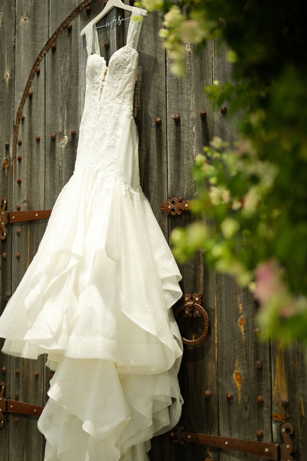 White lace mermaid wedding dress hanging on rustic wooden garden gate surrounded by green foliage and pink flowers.