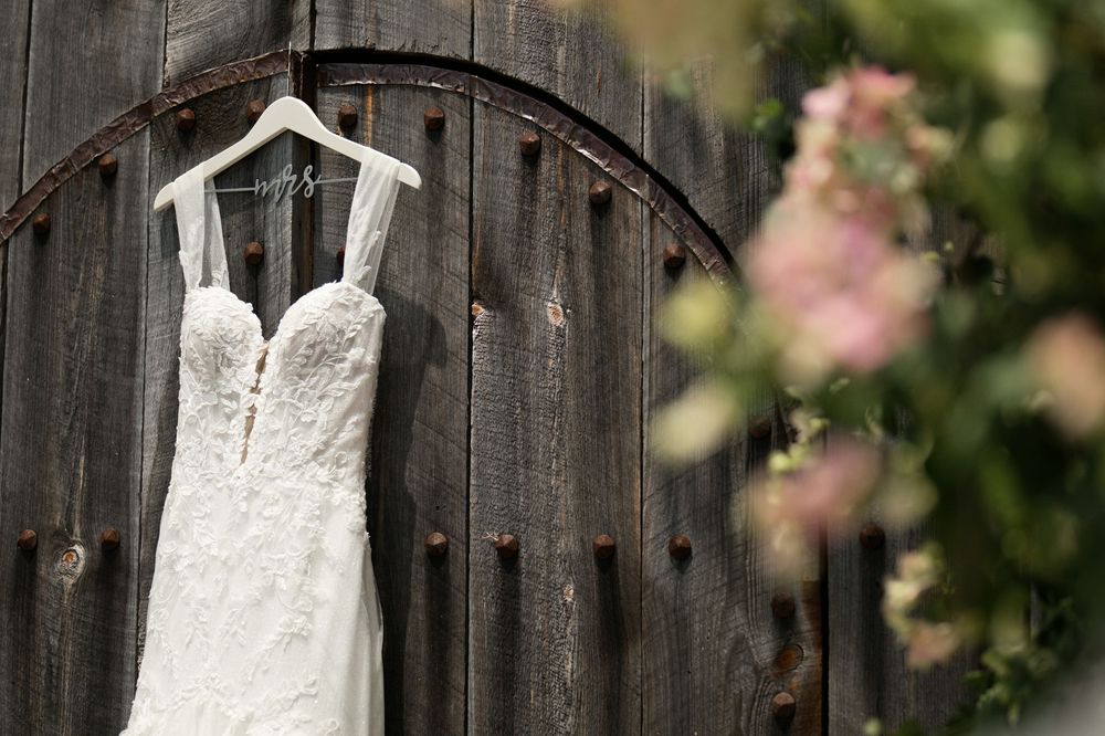 White lace wedding dress hanging on rustic wooden door with pink floral bokeh background.