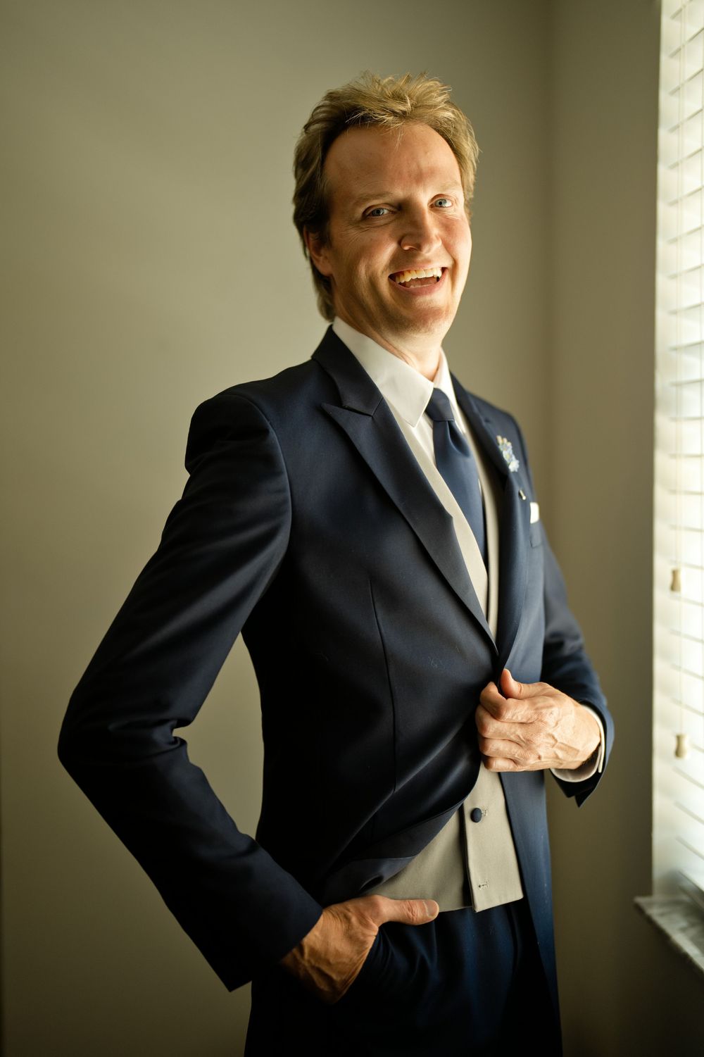 Smiling man in navy blue suit with tie and pocket square posing confidently near a window.