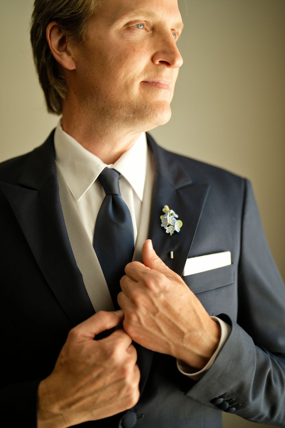 Groom in navy suit adjusting tie with boutonniere and white pocket square before wedding ceremony.