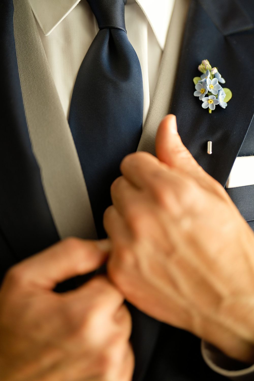A groom adjusts his navy tie on a suit jacket adorned with a delicate blue floral boutonniere.