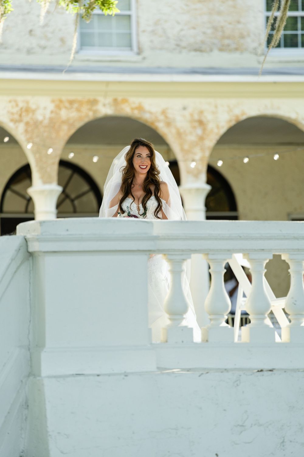 A smiling bride in a white wedding dress and veil stands on an elegant white balcony with arched columns behind her.